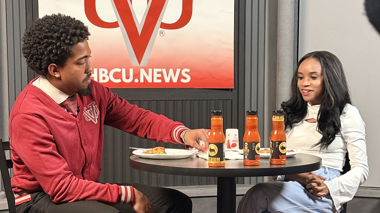 Mekhi McGainey and Jasmine Turner at a table with three sauce bottles in front of the HBCU.news backdrop